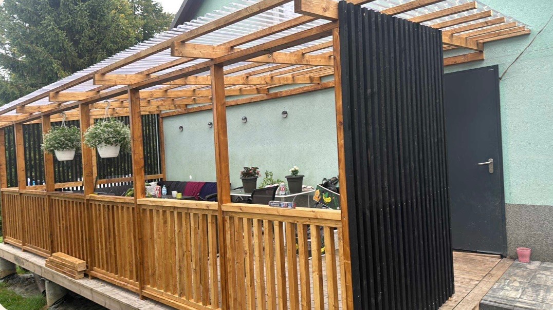 Drewniana pergola z przezroczystym zadaszeniem, balustradą i czarną, pionową osłoną. Wiszące donice z kwiatami, rośliny doniczkowe na stole, zielona ściana budynku w tle.