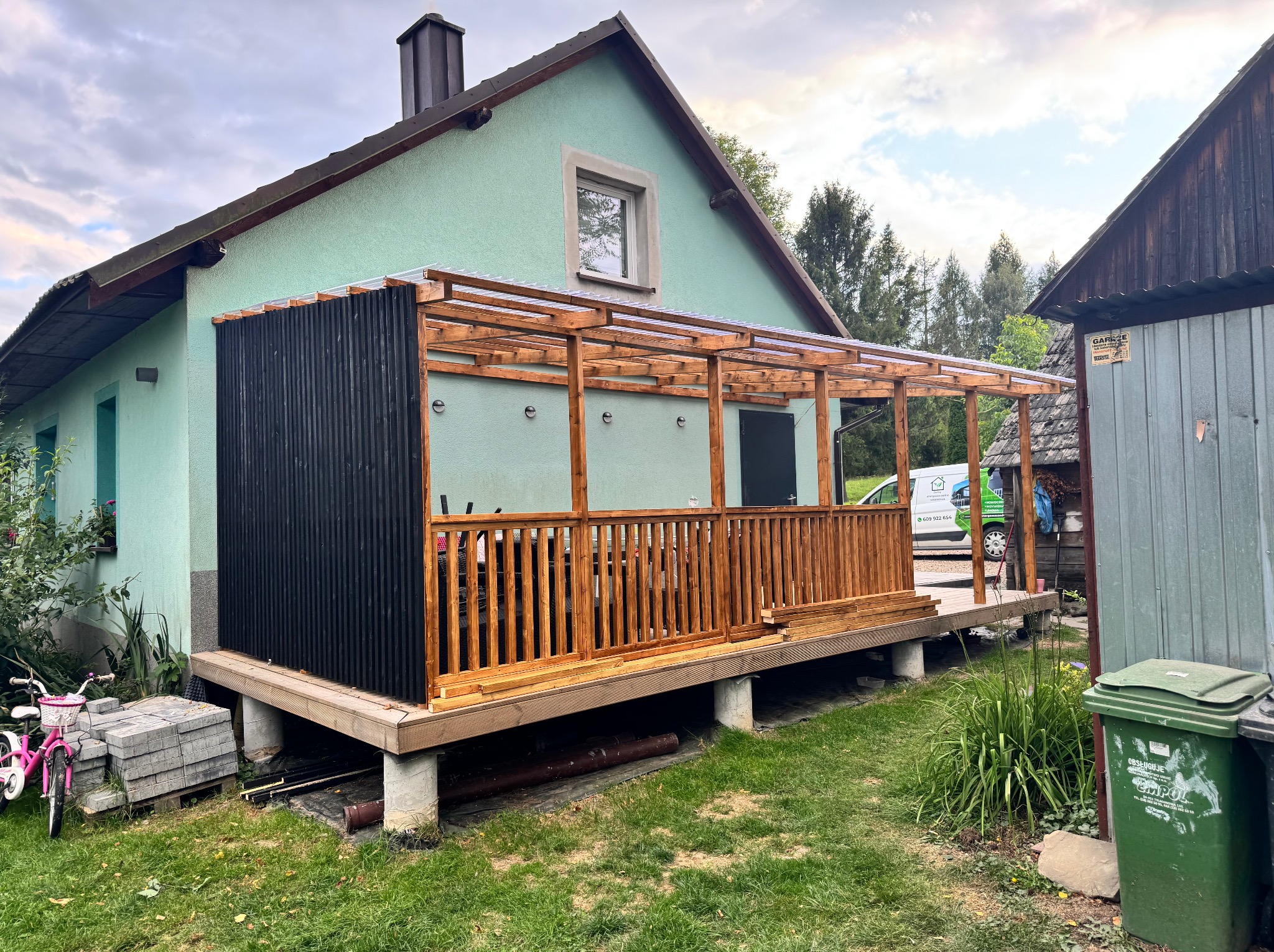 Nowa, drewniana konstrukcja z zadaszeniem, dobudowana do jasnego domu z zieloną elewacją. Widoczne elementy drewniane, betonowe podpory i częściowo czarna ściana.