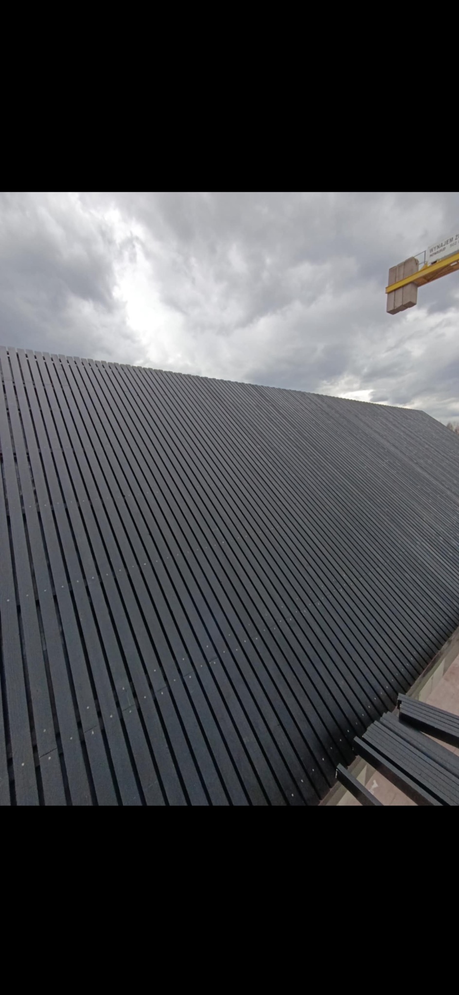 Ciemne, drewniane listwy elewacyjne ułożone pod kątem, tworzące nowoczesną fasadę budynku pod pochmurnym niebem. Widoczne mocowania i fragmenty konstrukcji.