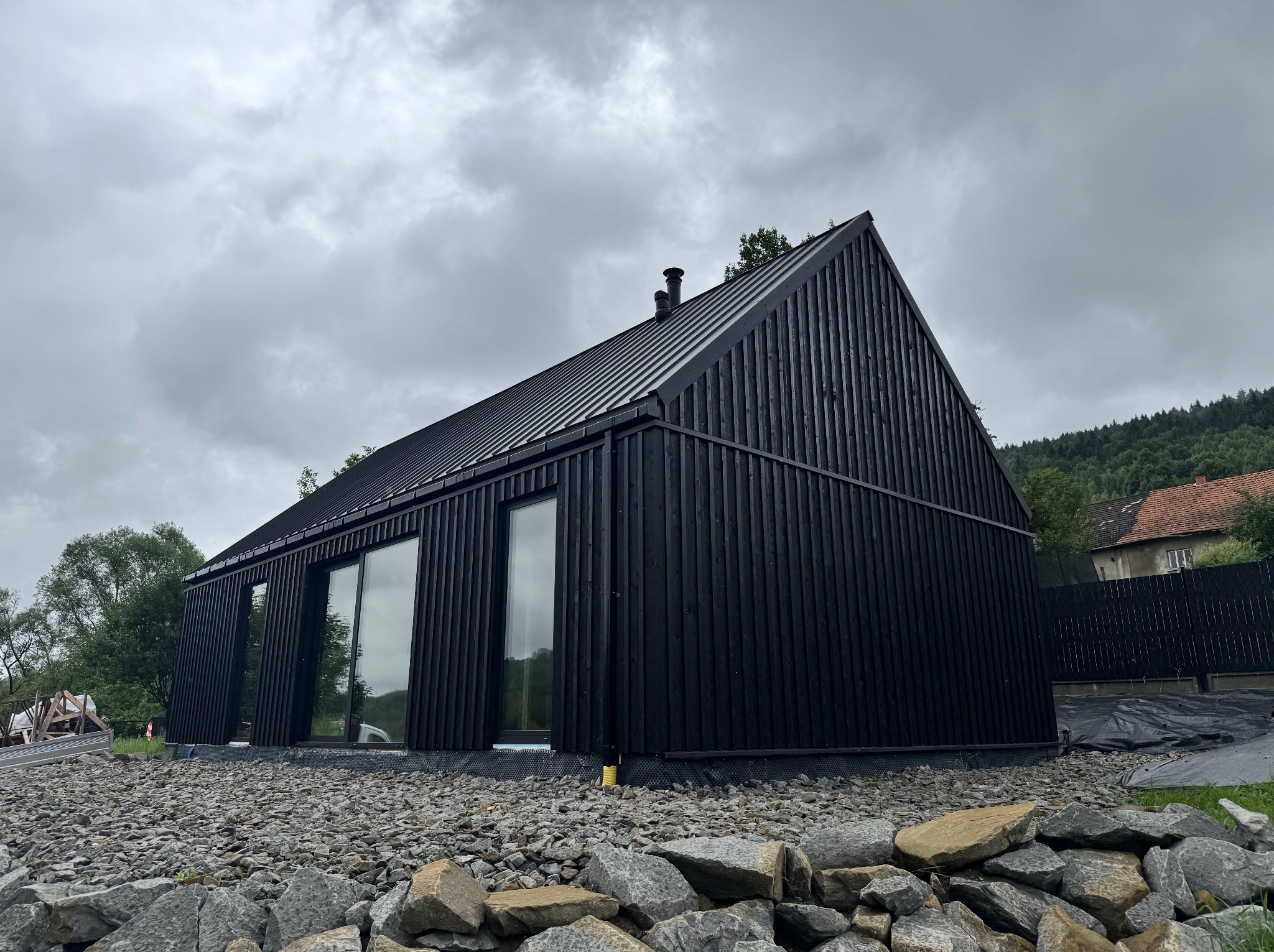 Czarny, nowoczesny dom z drewnianą elewacją i dużymi oknami, otoczony kamieniami. Widok na góry w tle, pochmurne niebo. Minimalistyczny design, ukończony projekt.