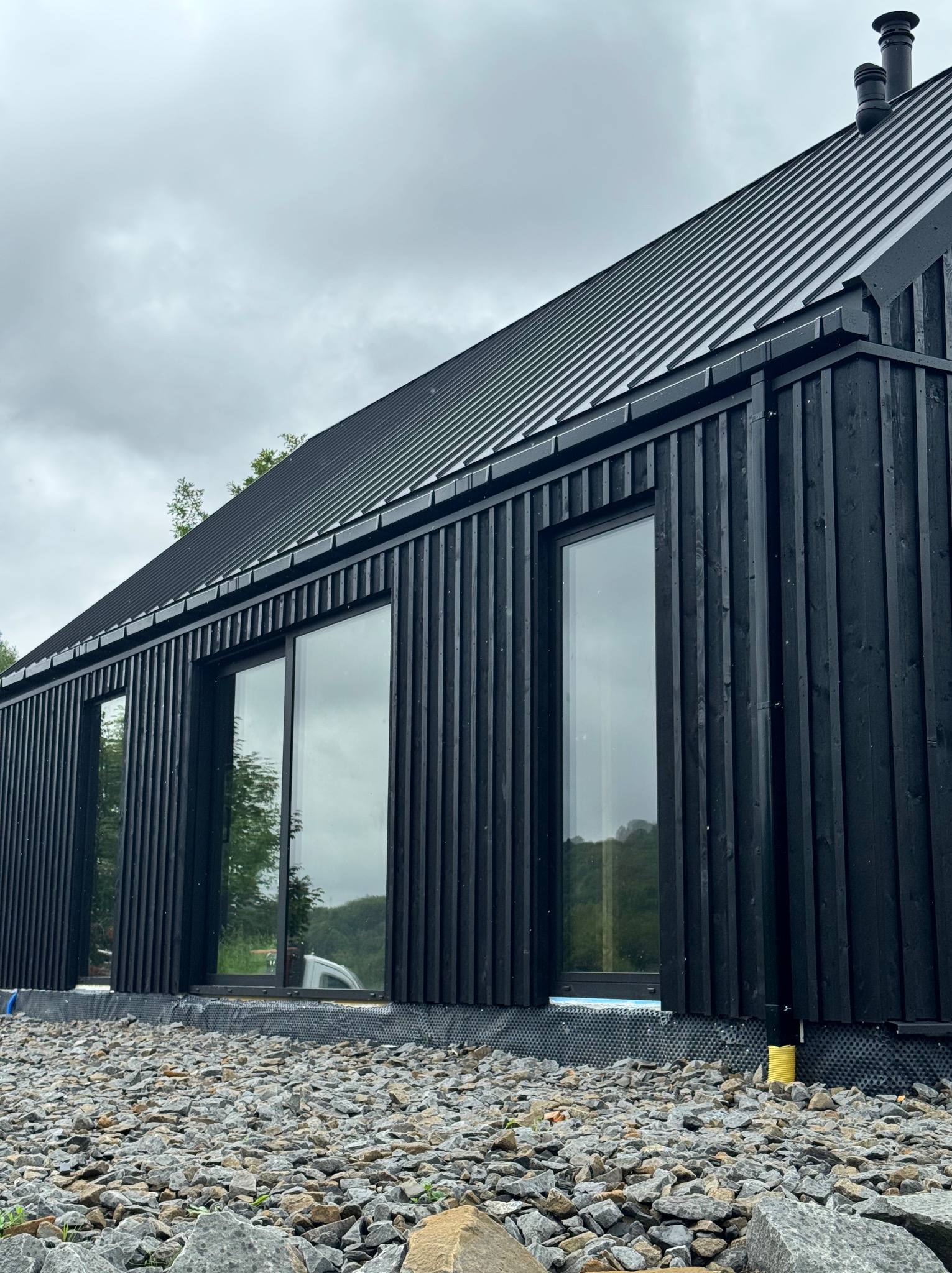 Nowoczesny dom z czarną, drewnianą elewacją i metalowym dachem, otoczony kamienistym terenem, widziany z perspektywy żabiej.