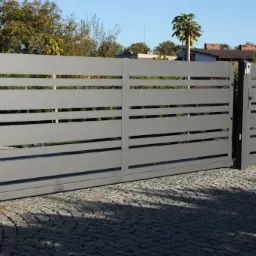 Nowoczesna, przesuwna brama wjazdowa w kolorze szarym z poziomymi szczelinami, skrzynka pocztowa po prawej stronie, kostka brukowa na podjeździe.