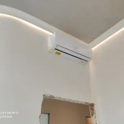 Nowy klimatyzator marki Gree, jeszcze w folii ochronnej, zamontowany w rogu pokoju z dekoracyjnym oświetleniem LED na suficie, widoczny otwór w ścianie po prawej stronie.