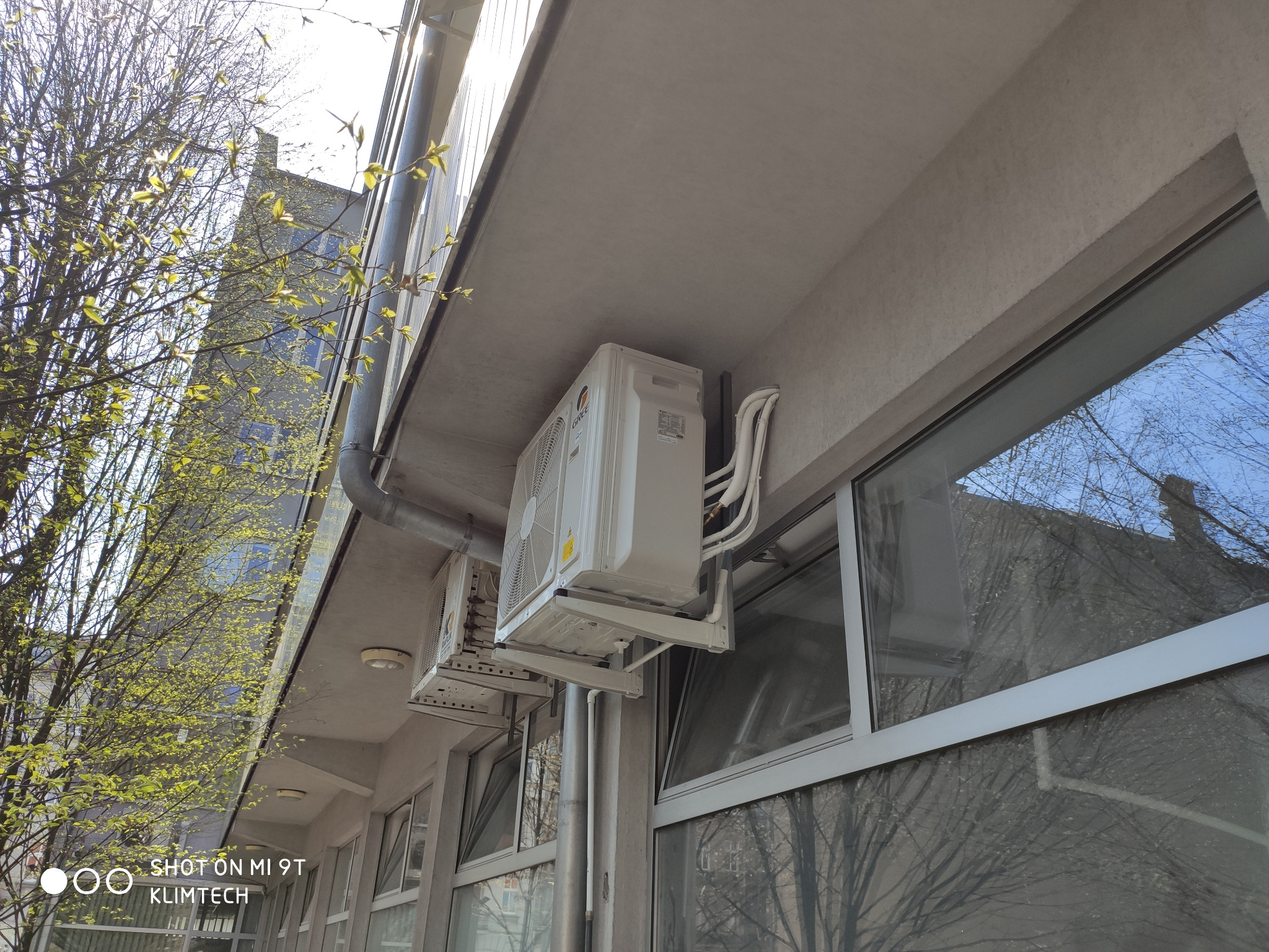 Zewnętrzne jednostki klimatyzacji zamontowane pod stropem budynku biurowego, widoczne białe rury i okna z odbijającym się drzewem.
