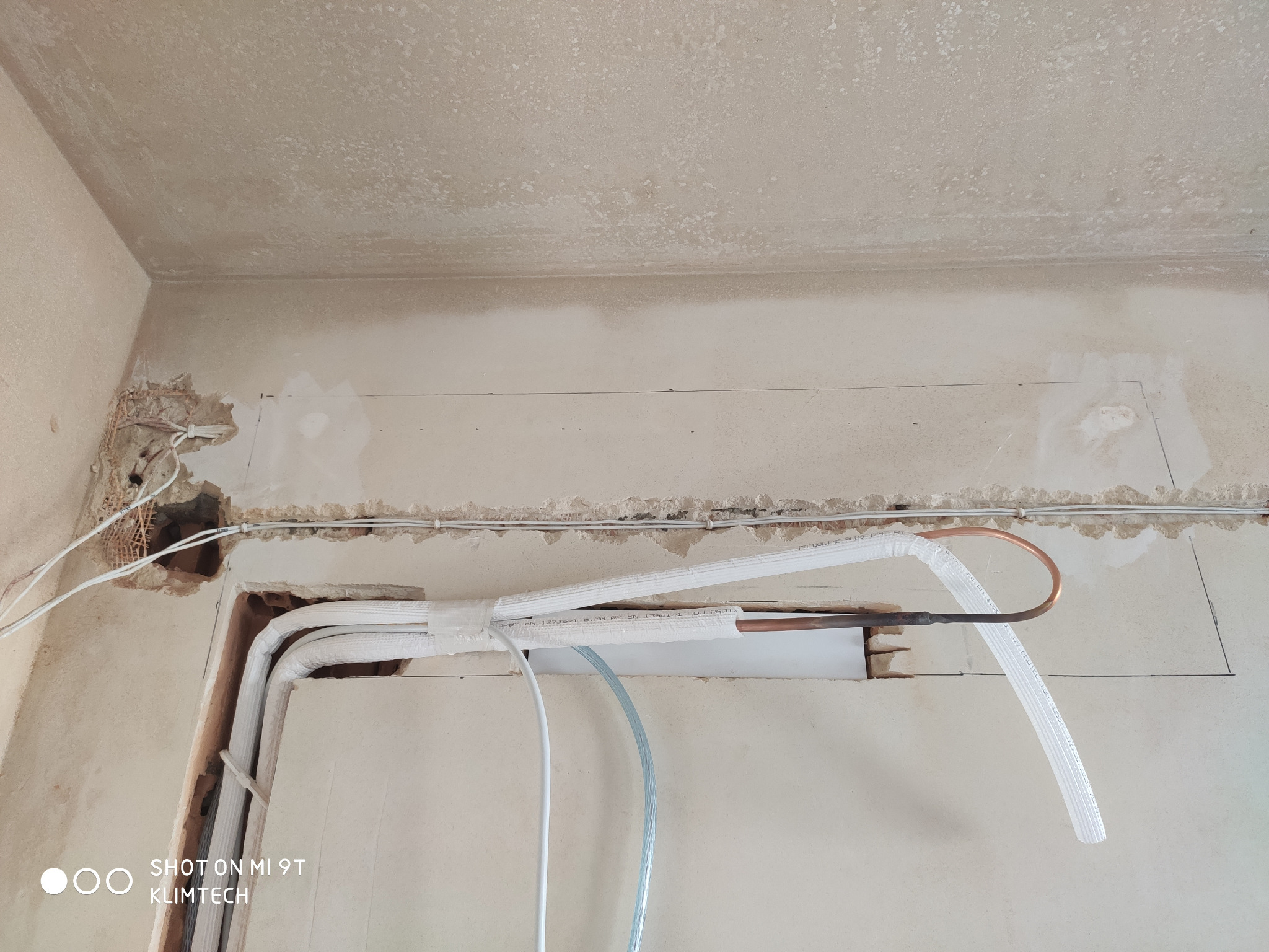 Instalacja rur miedzianych i przewodów w ścianie podczas montażu systemu chłodniczego, widoczne niedokończone wykończenie ściany i sufitu.