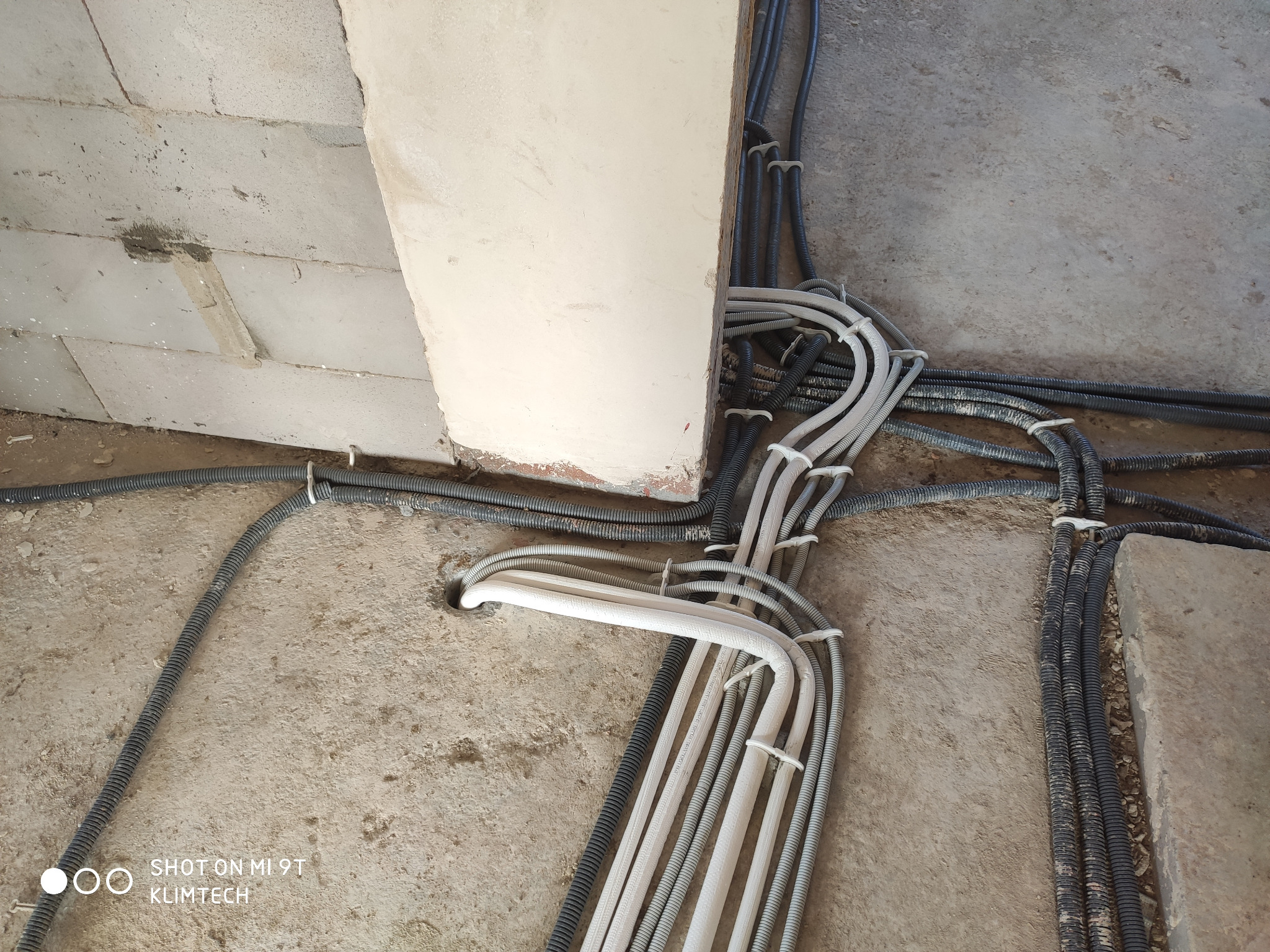 Instalacja elektryczna: wiązki rur karbowanych w kolorze białym i czarnym, przymocowane opaskami zaciskowymi, prowadzone po surowej podłodze i ścianie z betonu komórkowego.