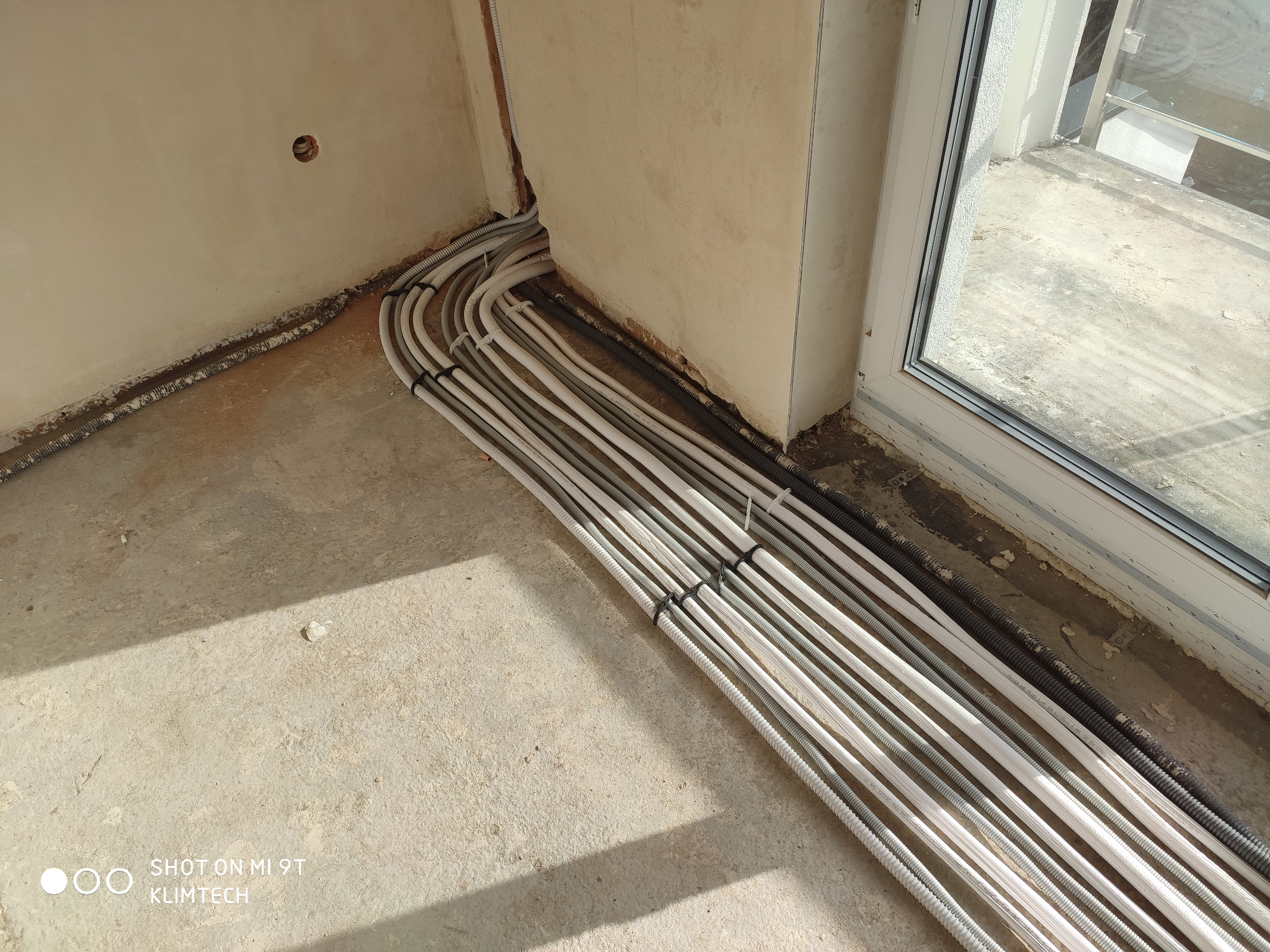 Rzędy białych i szarych rur instalacyjnych ułożone na surowym betonie pod ścianą z otworem, prowadzące w stronę okna balkonowego, zabezpieczone czarnymi opaskami zaciskowymi.