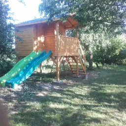 Drewniany domek dla dzieci z dwoma zjeżdżalniami w odcieniach zieleni i błękitu, otoczony zielonym trawnikiem i drzewami, widoczne betonowe podstawy i drewniane schody.