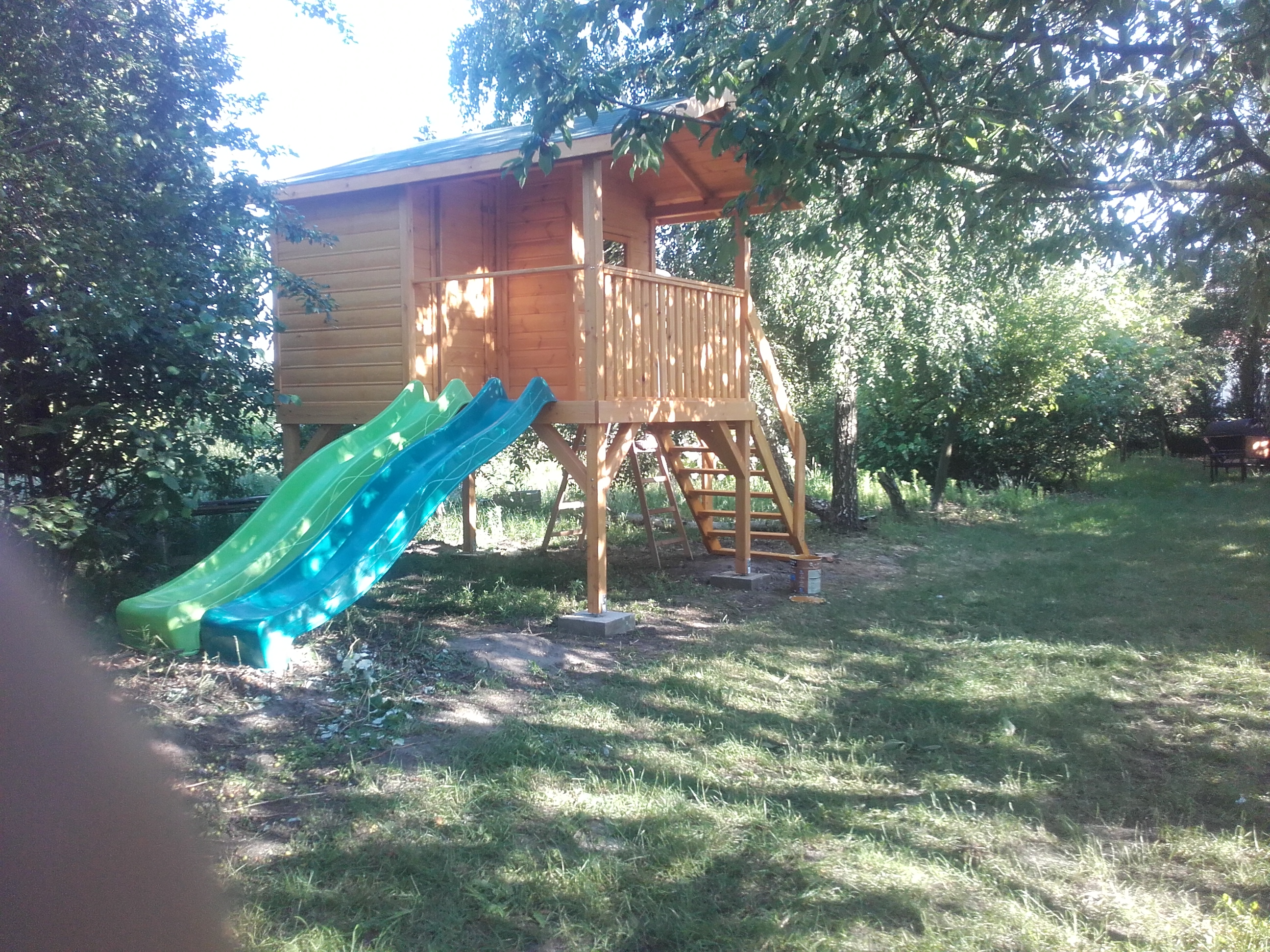 Drewniany domek dla dzieci z dwoma zjeżdżalniami w odcieniach zieleni i błękitu, otoczony zielonym trawnikiem i drzewami, widoczne betonowe podstawy i drewniane schody.