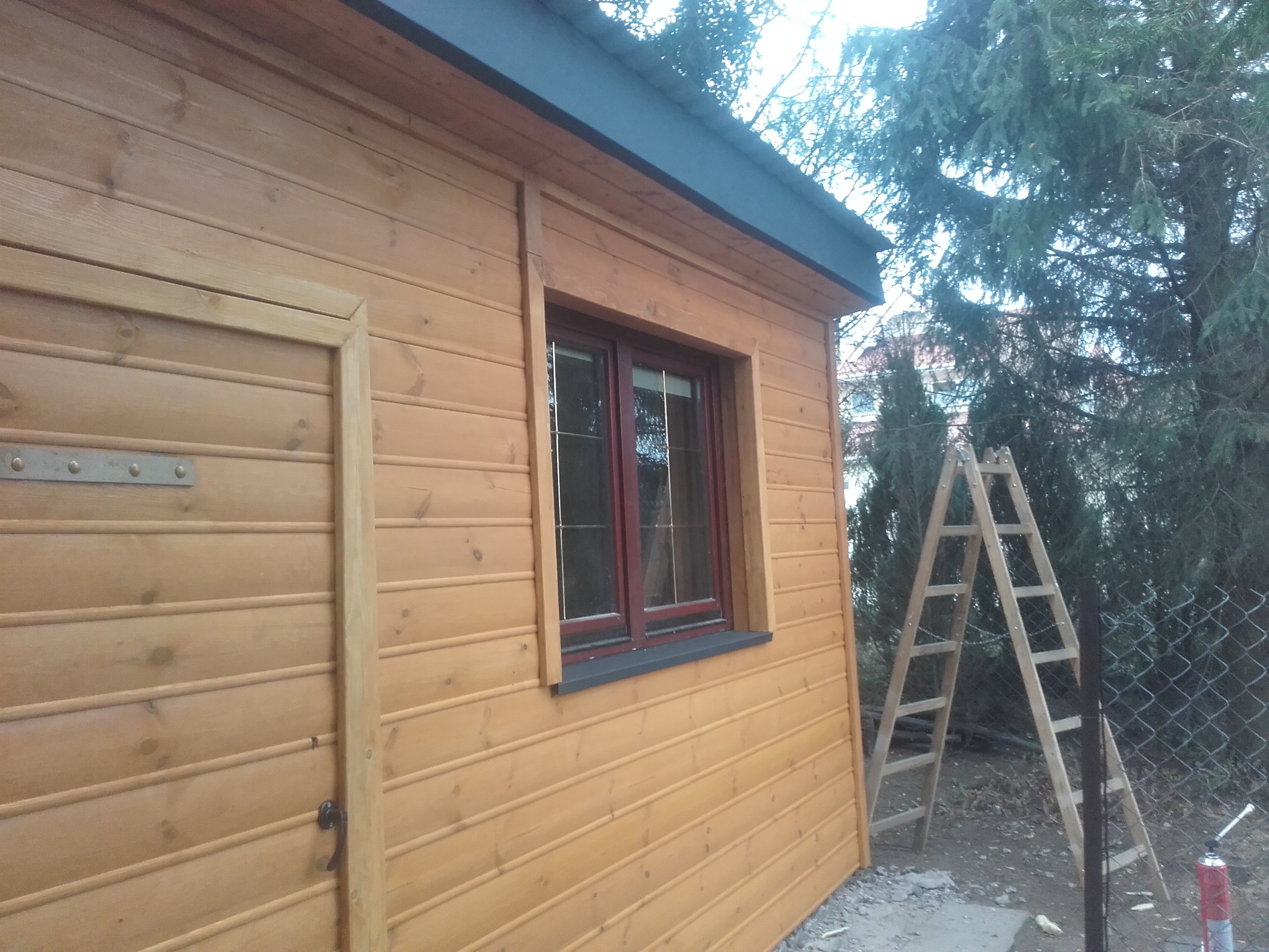 Świeżo wybudowana, drewniana altana z oknem w czerwonej ramie i drzwiami z widocznymi okuciami, częściowo pomalowana, z drabiną opartą o ścianę i butlą z gazem w tle.