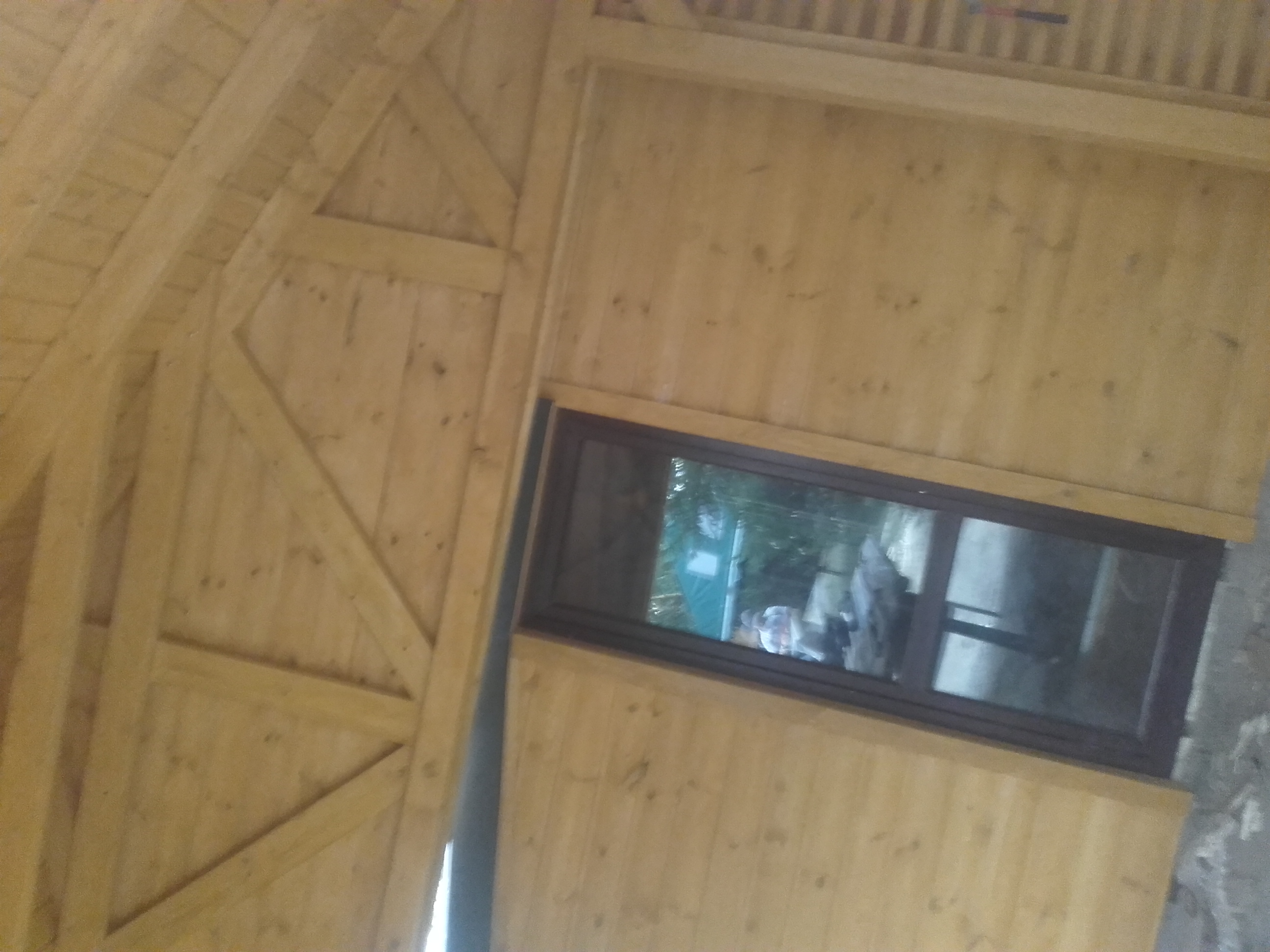 Wnętrze drewnianej konstrukcji z widocznym oknem, ukazującym fragment otoczenia zewnętrznego z zielenią i postaciami w oddali.