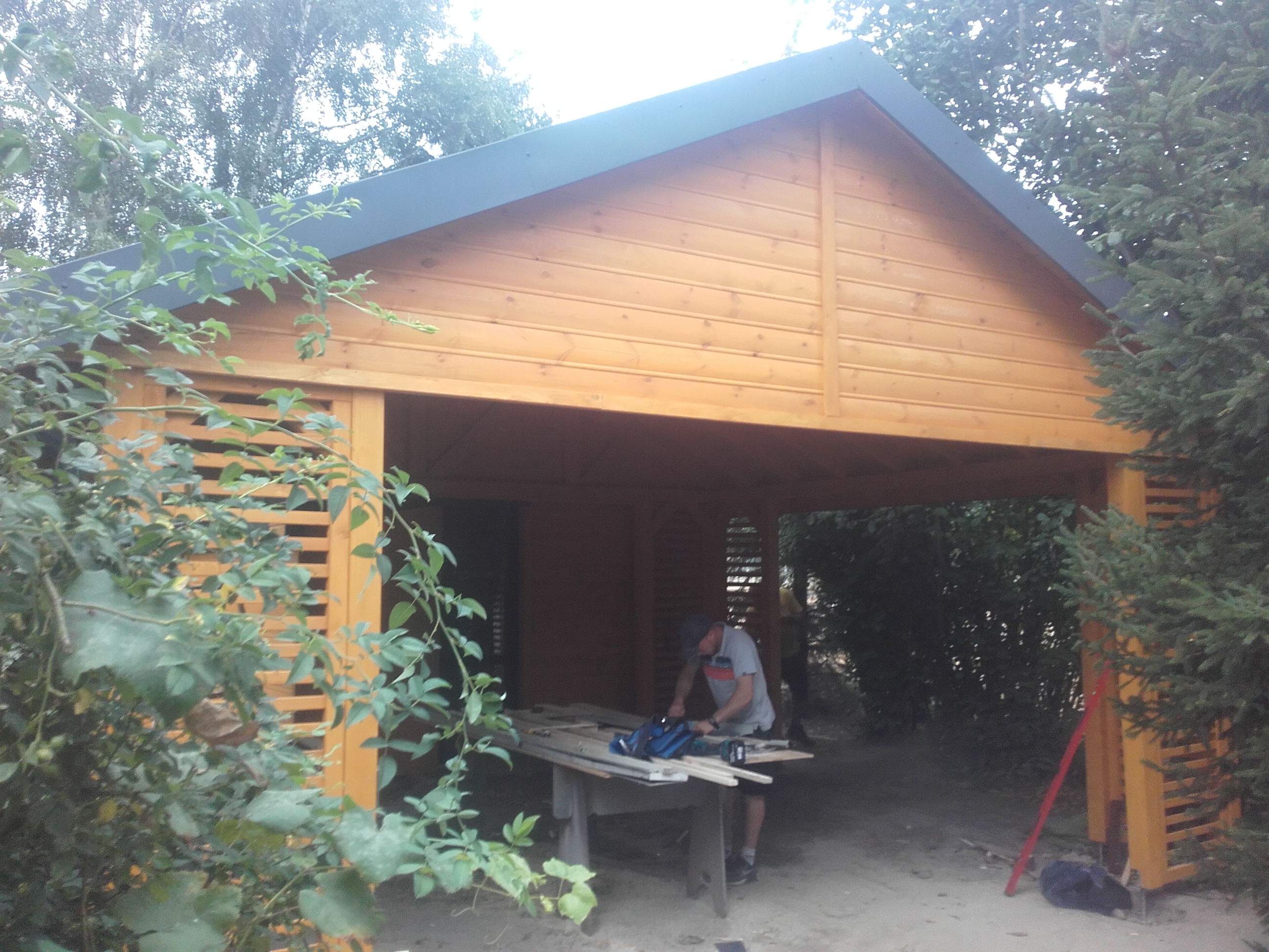 Budowa drewnianej altany z ażurowymi ścianami bocznymi; widoczny mężczyzna przycinający deski na stole roboczym, narzędzia i elementy konstrukcyjne w tle.