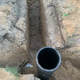 Głęboki wykop w ziemi z widoczną czarną rurą z tworzywa sztucznego, fragment folii i trawy przy krawędzi wykopu.