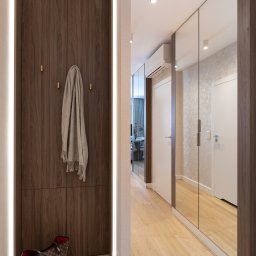 Lignum Design S.C. Monika Jakubowska Zbigniew Krupa - Wnętrze przedpokoju z zabudowaną szafą z forniru orzechowego z oświetleniem LED po bokach, w tle szafa z lustrzanymi drzwiami i drewniana podłoga.
