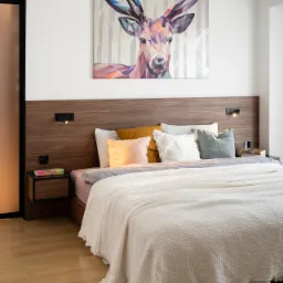 Nowoczesna sypialnia z drewnianym wezgłowiem, nocną szafką z kolorowym akcentem i obrazem geometrycznego jelenia nad łóżkiem.