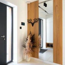 Nowoczesny przedpokój z drewnianymi panelami, lustrem, dekoracją ścienną w kształcie głowy jelenia, pampasową trawą w wazonie i szarą wycieraczką przy wejściu.