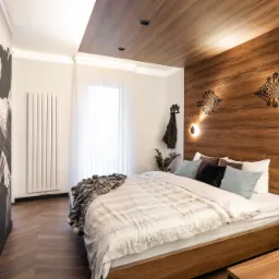 Nowoczesna sypialnia z drewnianymi panelami ściennymi i sufitowymi, ozdobiona czarnymi, etnicznymi wzorami i minimalistycznymi lampami ściennymi, łóżko z narzutą w odcieniach szarości i beżu...