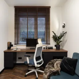 Biuro domowe z drewnianym blatem biurka, czarnymi szafkami, ergonomicznym krzesłem biurowym, drewnianymi żaluzjami i wzorem ludowym na ścianie.