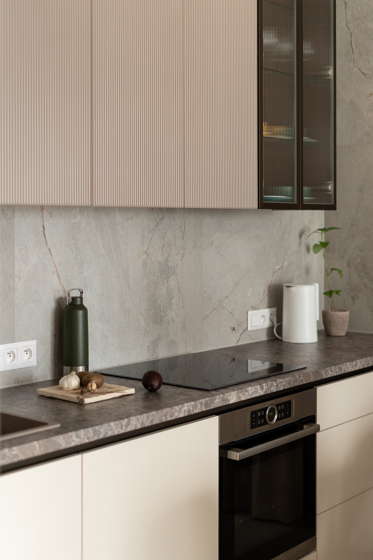 Nowoczesna kuchnia z beżowymi szafkami górnymi o ryflowanych frontach i szklanymi witrynami w ciemnych ramach, szary blat z płyty imitującej kamień, indukcja, piekarnik, akcesoria kuchenne.