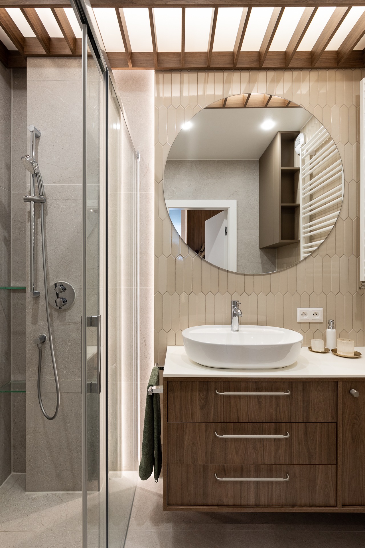 Nowoczesna łazienka z kabiną prysznicową, okrągłym lustrem i drewnianą szafką pod umywalką z trzema szufladami, oświetlenie LED za lustrem i w kabinie.