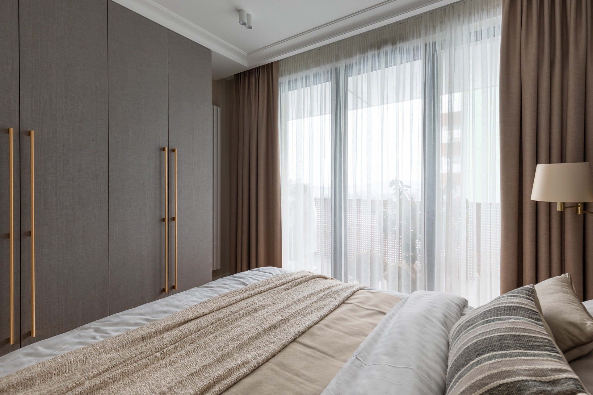 Szafa na wymiar w sypialni z długimi, złotymi uchwytami, dopasowana kolorystycznie do zasłon i narzuty na łóżku.