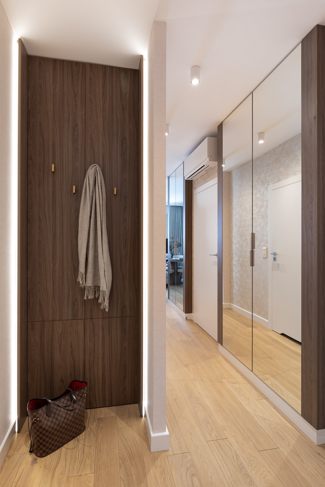 Wnętrze przedpokoju z zabudowaną szafą z forniru orzechowego z oświetleniem LED po bokach, w tle szafa z lustrzanymi drzwiami i drewniana podłoga.