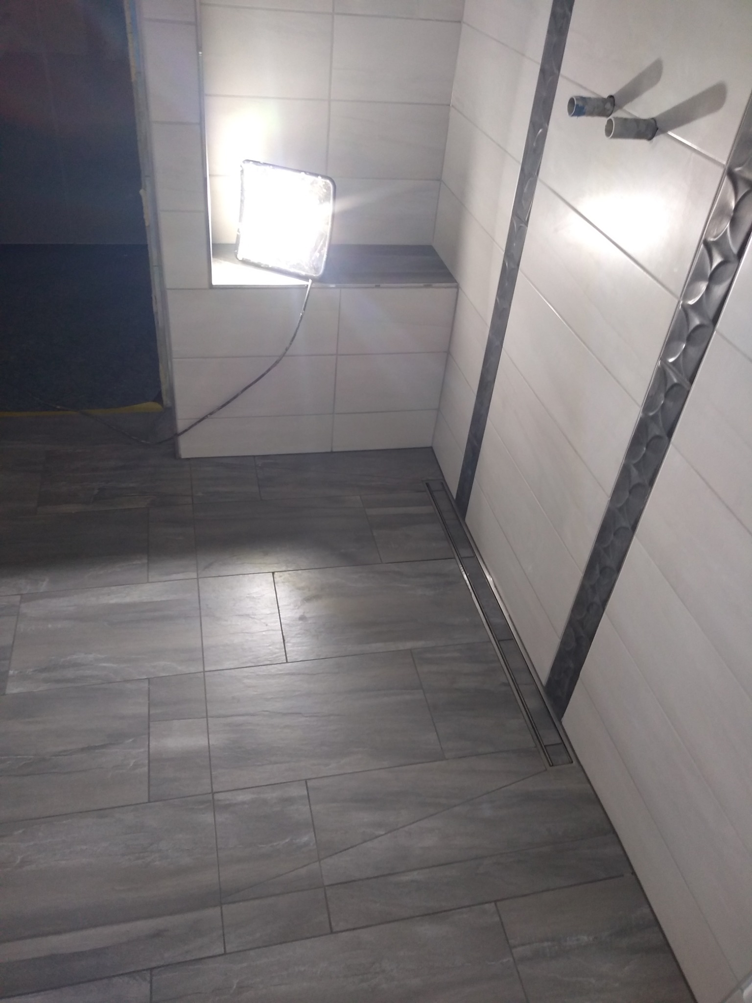 Wykończona łazienka z szarymi płytkami podłogowymi i białymi płytkami ściennymi z dekoracyjnymi listwami. Widoczne rury instalacji wodnej wystające ze ściany oraz lampa robocza oświetlająca wnętrze.