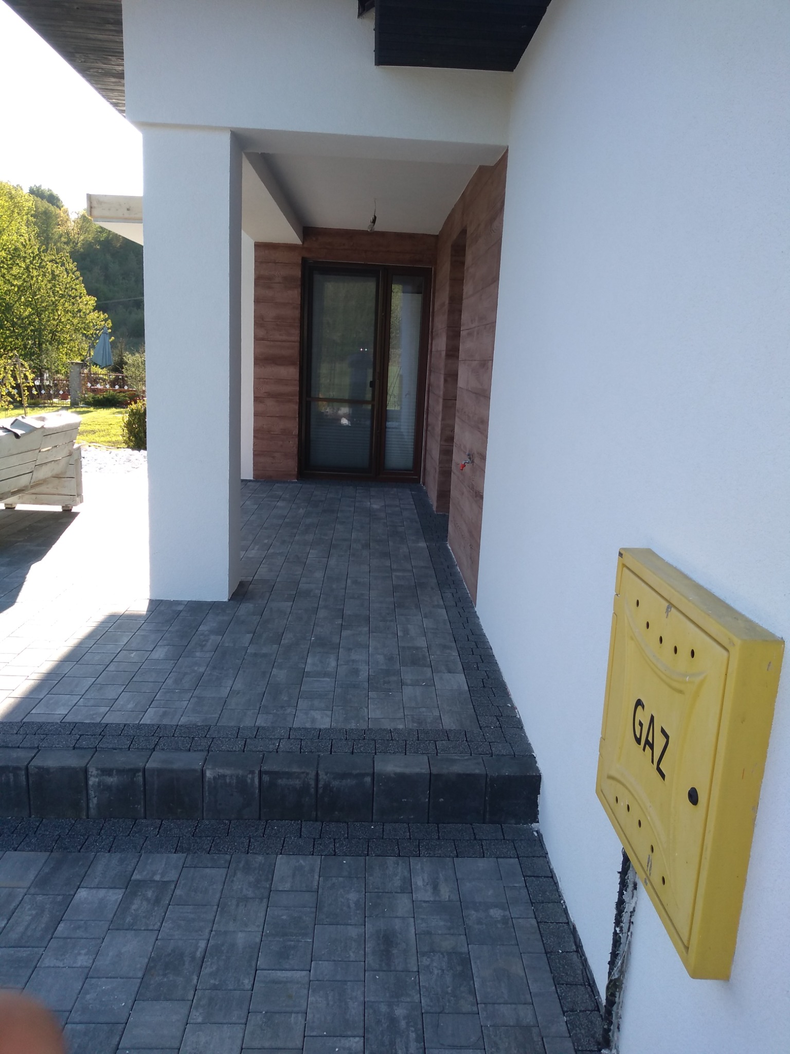 Wejście do domu z wyłożonymi kostką brukową schodami i chodnikiem, z widoczną skrzynką gazową na ścianie budynku.