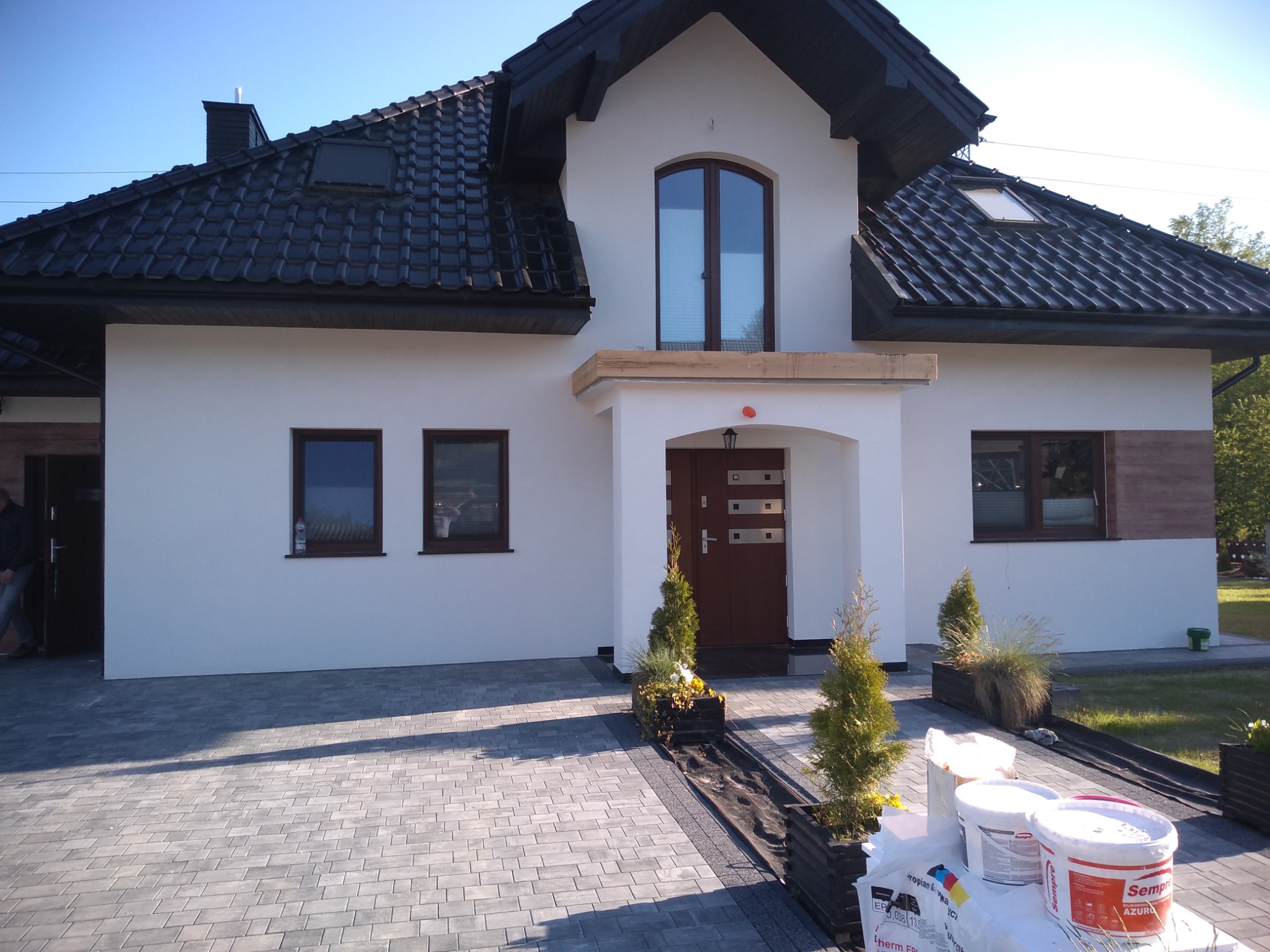 Nowoczesny dom jednorodzinny z białymi ścianami i ciemną dachówką, widoczne puszki z farbą i worki z materiałami budowlanymi przed wejściem, sugerujące prace wykończeniowe.
