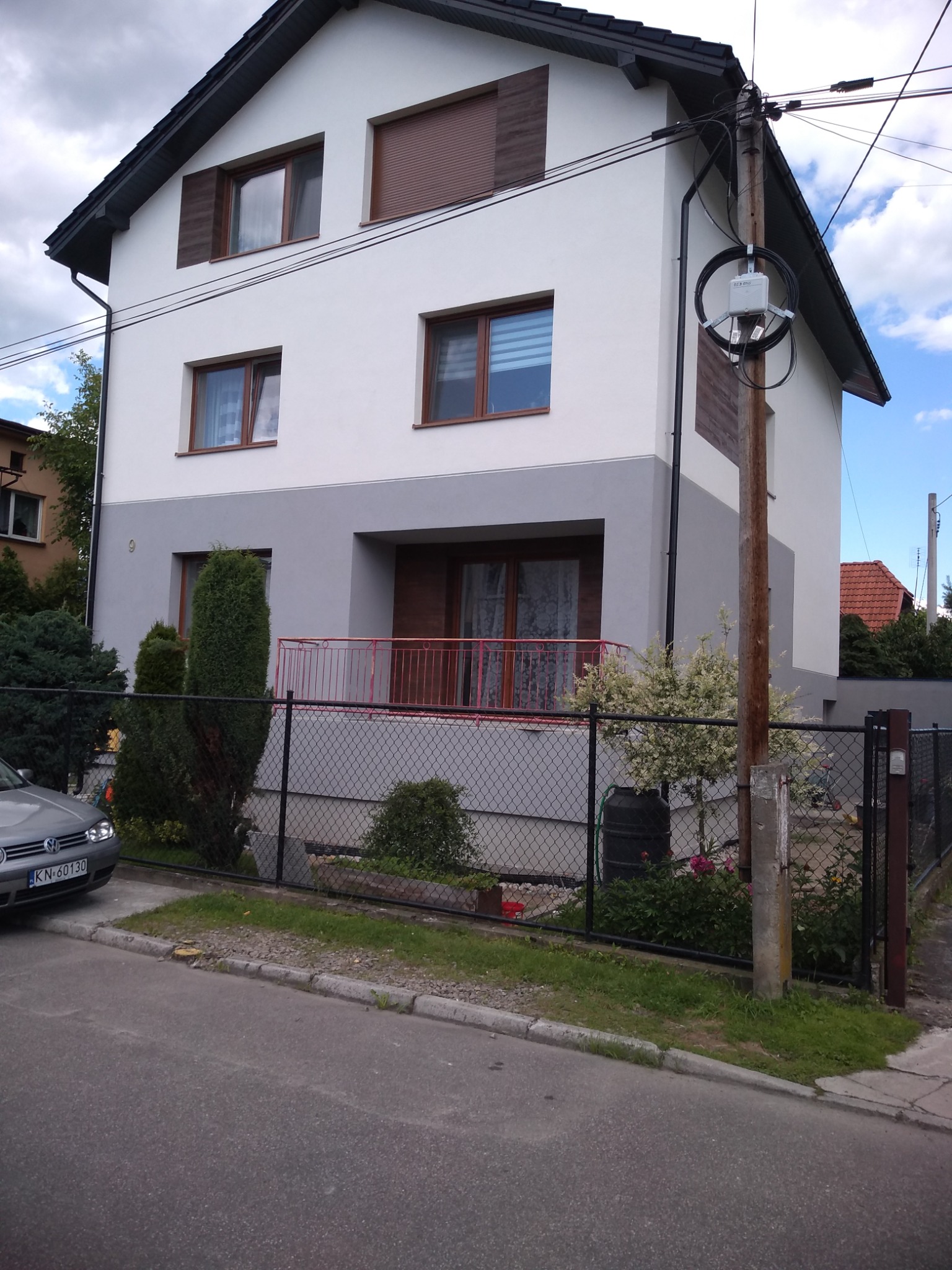 Dwukondygnacyjny dom z białymi i szarymi ścianami, brązowymi oknami i roletami, z czarnym ogrodzeniem i zielenią w ogrodzie, widoczny słup energetyczny z okablowaniem.