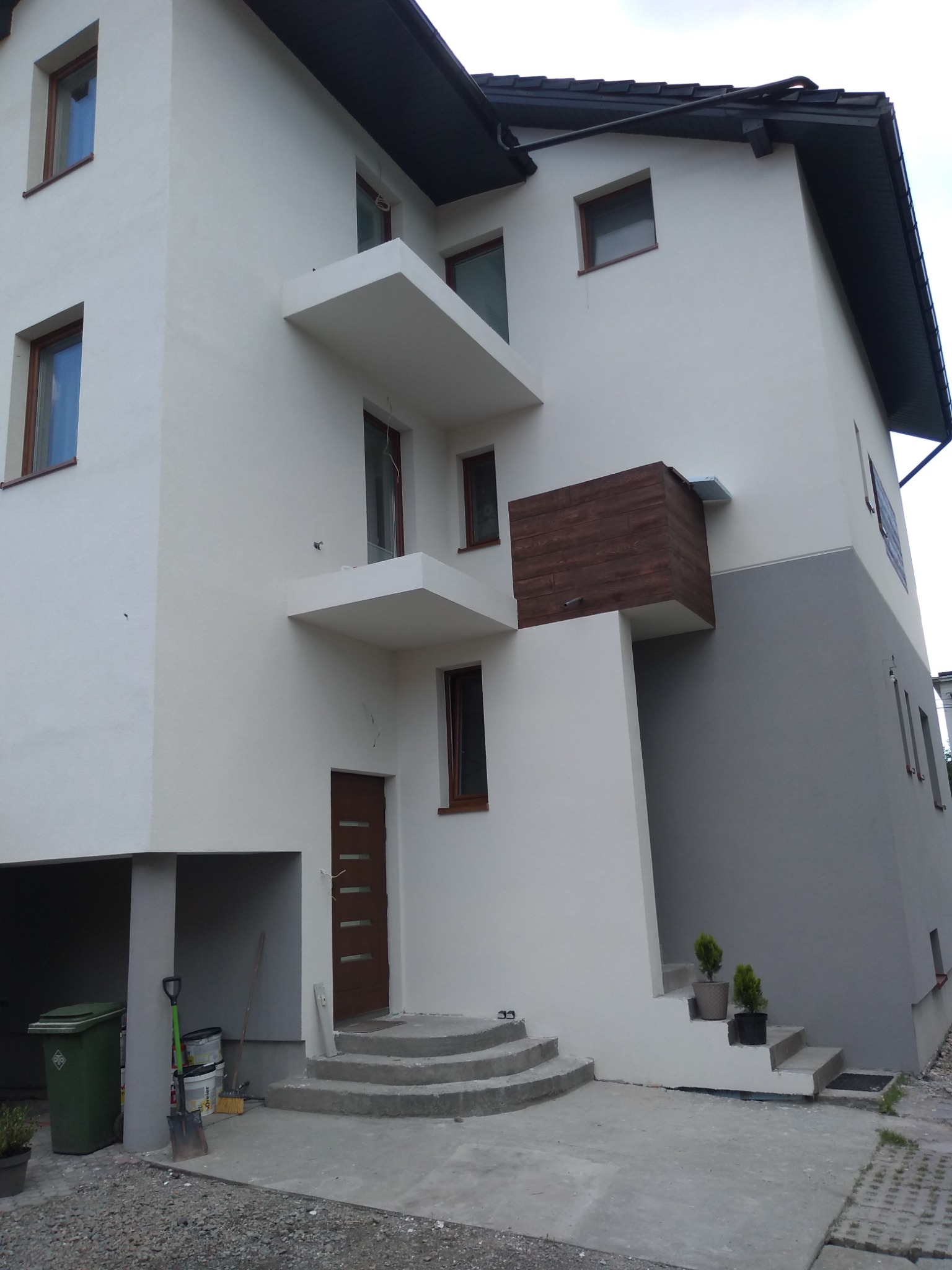 Nowoczesny dom jednorodzinny po remoncie elewacji, z białymi ścianami na górze i szarymi na dole, drewnianym elementem dekoracyjnym, balkonami, schodami wejściowymi i widocznymi narzędziami...