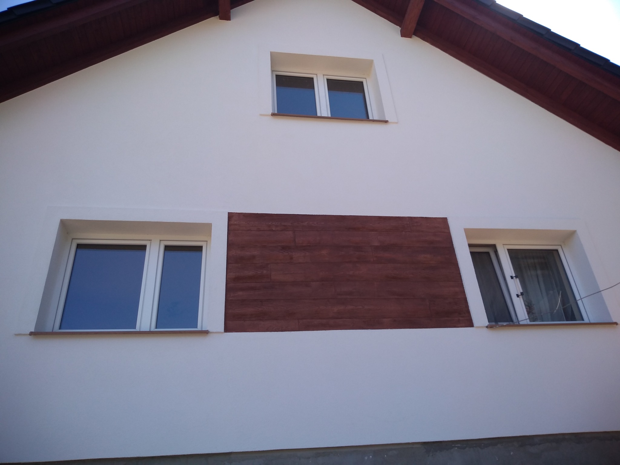 Biała fasada domu jednorodzinnego z trzema oknami i dekoracyjnym panelem z imitacji drewna, widok z dołu