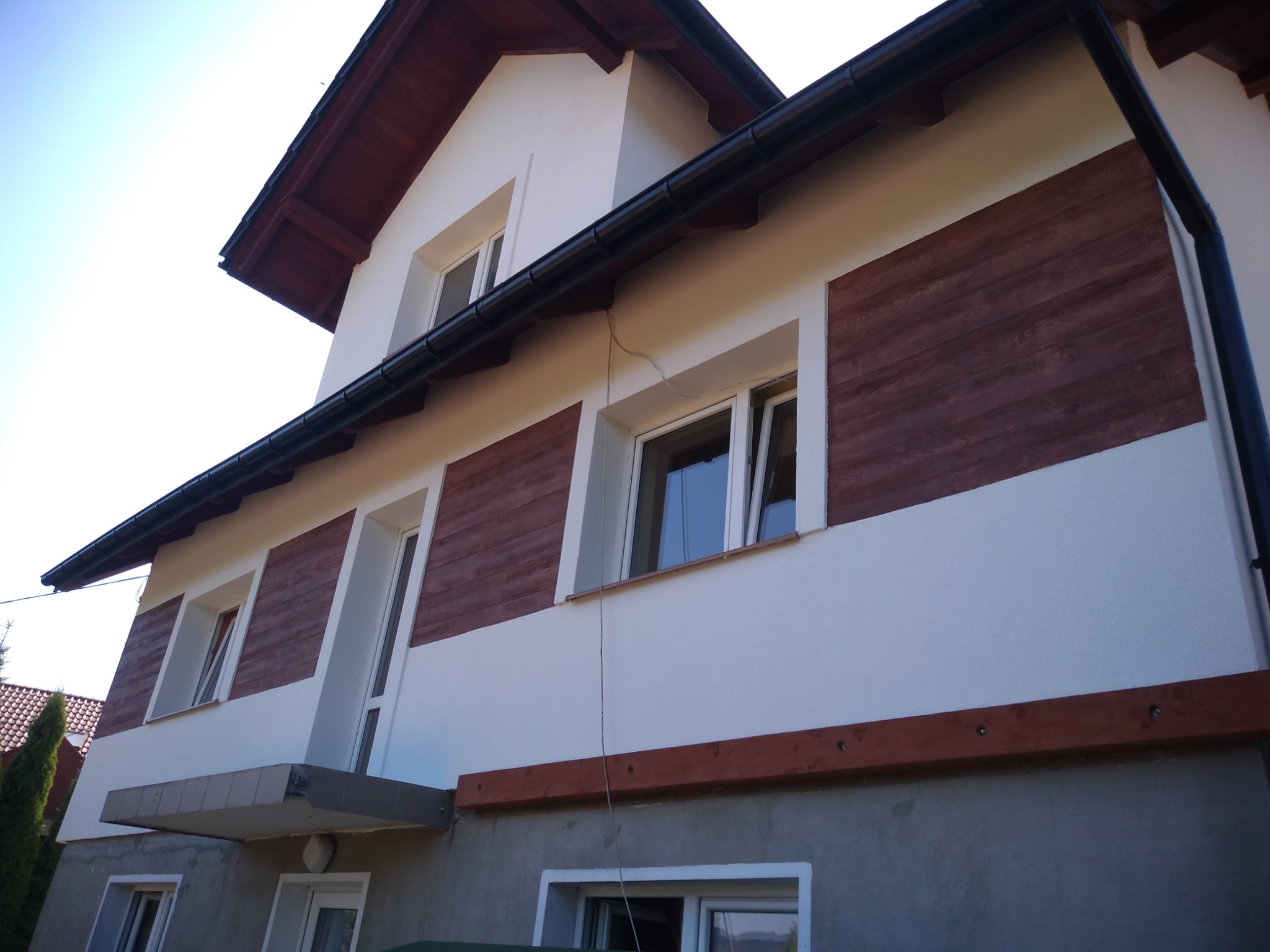 Dwupiętrowy dom z białymi ścianami i poziomymi pasami z desek w kolorze ciemnego drewna, widoczny z dołu pod kątem, z czarną rynną dachową i częściowo widocznym dachem z ciemnej dachówki.