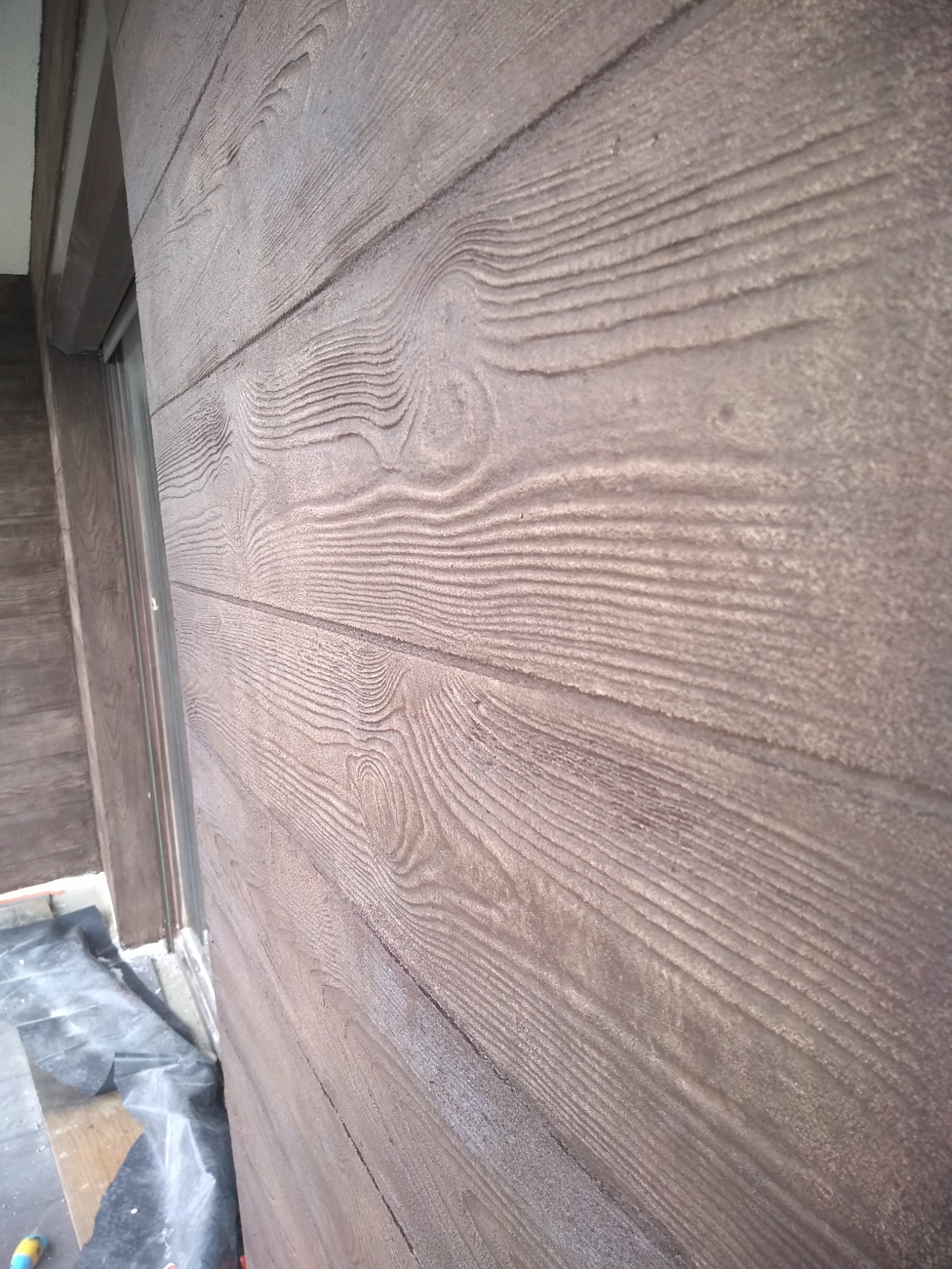 Ocieplona ściana zewnętrzna budynku z brązową strukturą imitującą drewno, widoczna przy oknie z zabezpieczającą folią na podłożu.