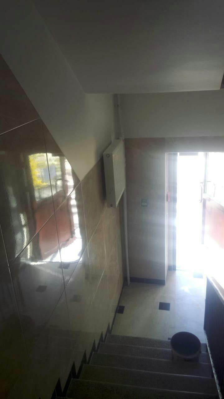 Klatka schodowa w budynku, wyłożona brązowymi, błyszczącymi płytkami ceramicznymi. Widoczny grzejnik na ścianie, schody z czarnymi akcentami i otwarte drzwi na końcu korytarza. Na schodach stoi...