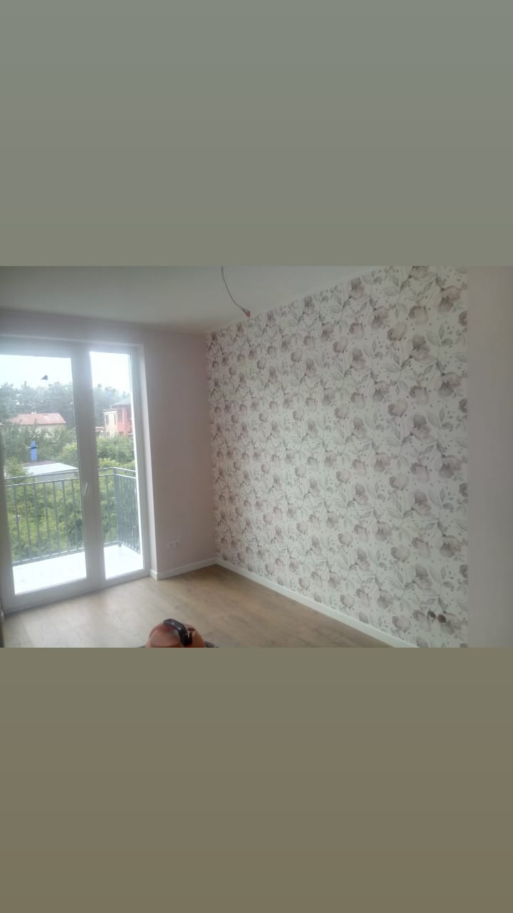 Świeżo wytapetowany pokój z balkonem; ściana pokryta tapetą w delikatne, szaro-białe kwiaty, jasna drewniana podłoga, białe drzwi balkonowe, widok na zieleń za oknem, wystający kabel ze ściany.