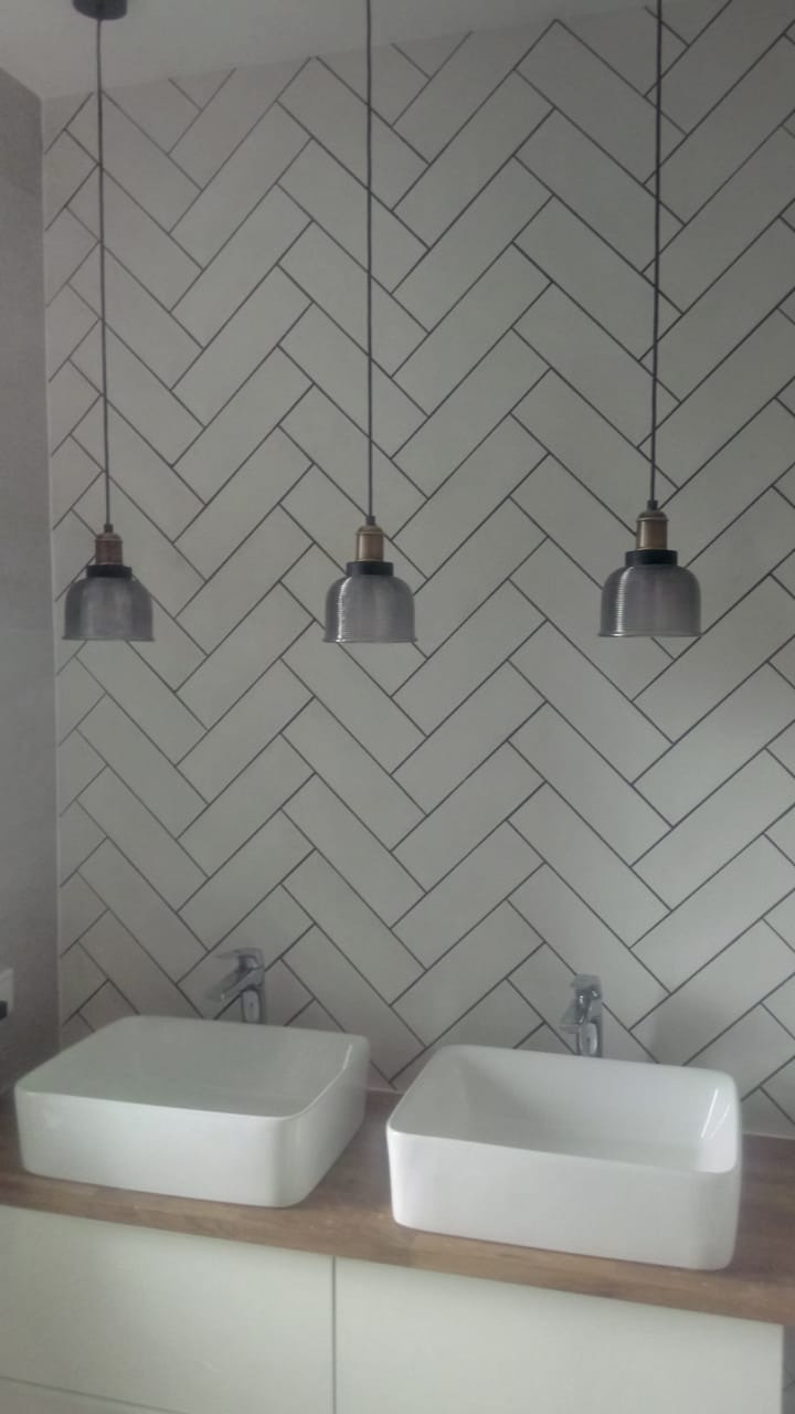 Nowoczesna łazienka z dwiema kwadratowymi umywalkami nablatowymi, bateriami chromowanymi i ścianą wyłożoną białymi płytkami ułożonymi w jodełkę, oświetlona trzema wiszącymi lampami w stylu...