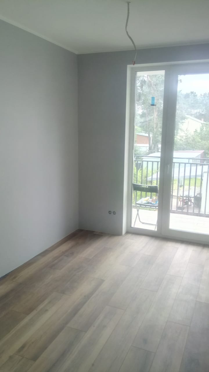 Świeżo pomalowane na szaro pomieszczenie z drewnianą podłogą, balkonem i wystającym kablem ze ściany.