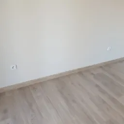 Pusty pokój po remoncie: jasne panele podłogowe, kremowe ściany z dwoma gniazdkami elektrycznymi i przypodłogowa listwa dekoracyjna w kolorze drewna.