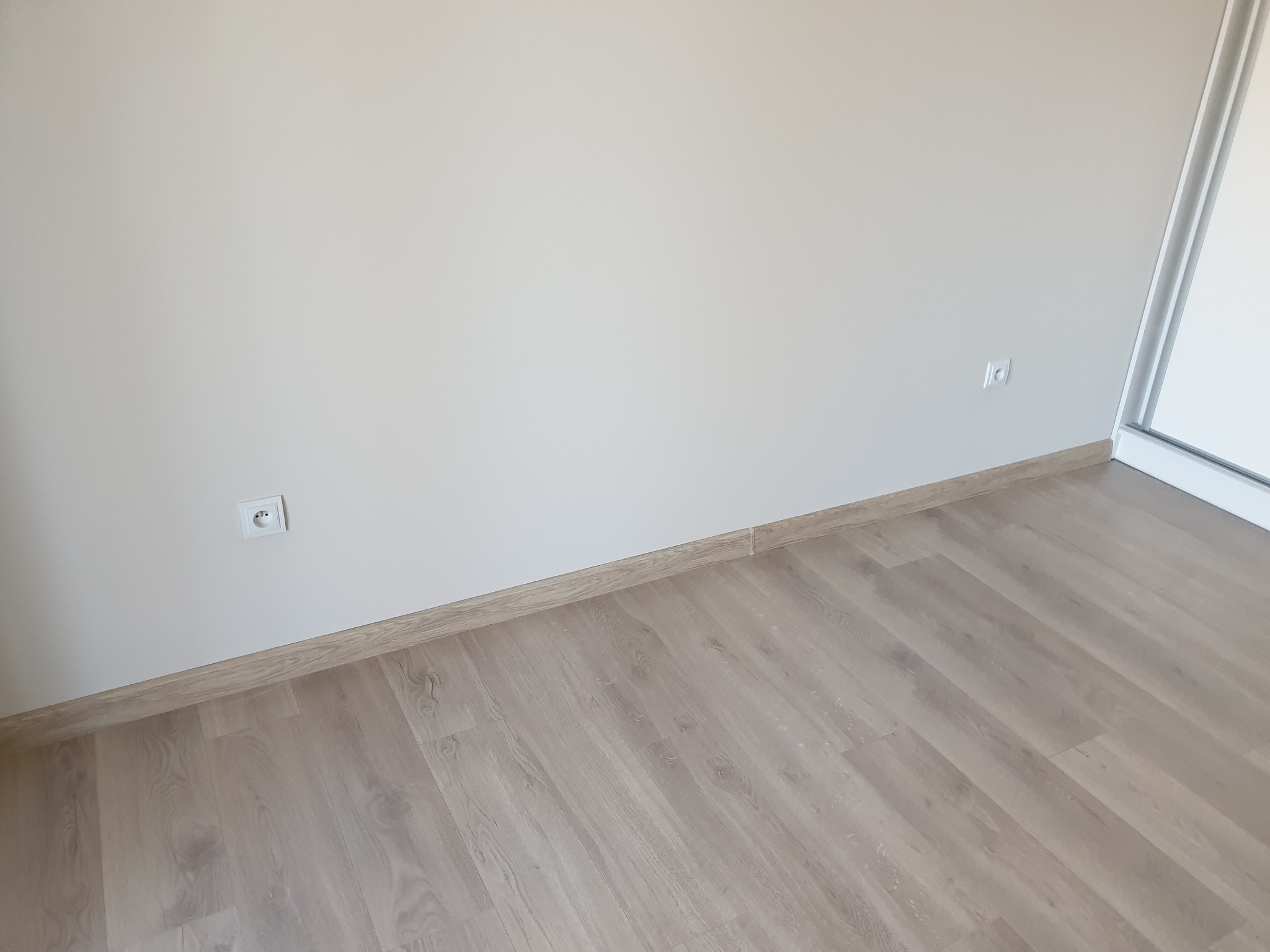 Pusty pokój po remoncie: jasne panele podłogowe, kremowe ściany z dwoma gniazdkami elektrycznymi i przypodłogowa listwa dekoracyjna w kolorze drewna.