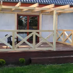 W trakcie budowy: drewniana konstrukcja z balustradą w kształcie krzyży, zadaszenie z belek, widoczny fragment tarasu i schody, w tle ściana budynku z oknami i drzwiami.