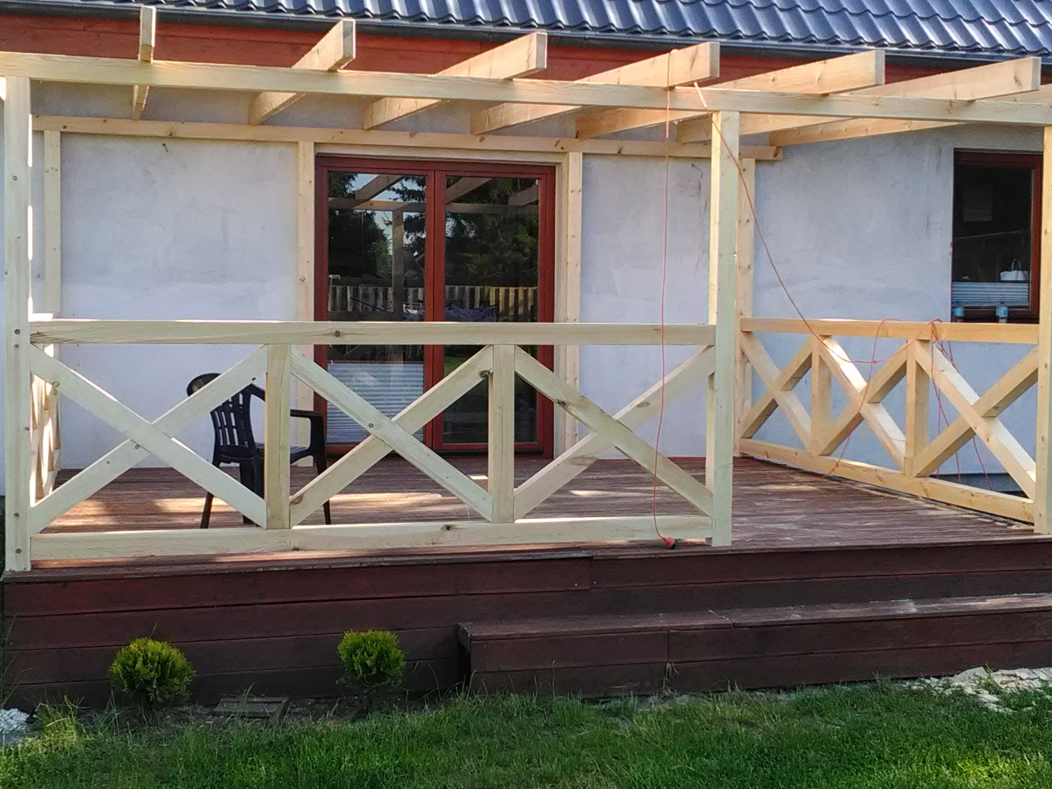 W trakcie budowy: drewniana konstrukcja z balustradą w kształcie krzyży, zadaszenie z belek, widoczny fragment tarasu i schody, w tle ściana budynku z oknami i drzwiami.