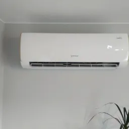 Biała klimatyzacja Rotenso typu split z wyświetlaczem temperatury 24 stopnie Celsjusza, zamontowana na szarej ścianie w rogu pokoju, z widoczną rośliną doniczkową w prawym dolnym rogu.