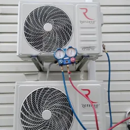 Dwa białe klimatyzatory typu split marki Rotenso zamontowane jeden nad drugim na elewacji budynku, podłączone do manometrów serwisowych za pomocą czerwonych i niebieskich węży.