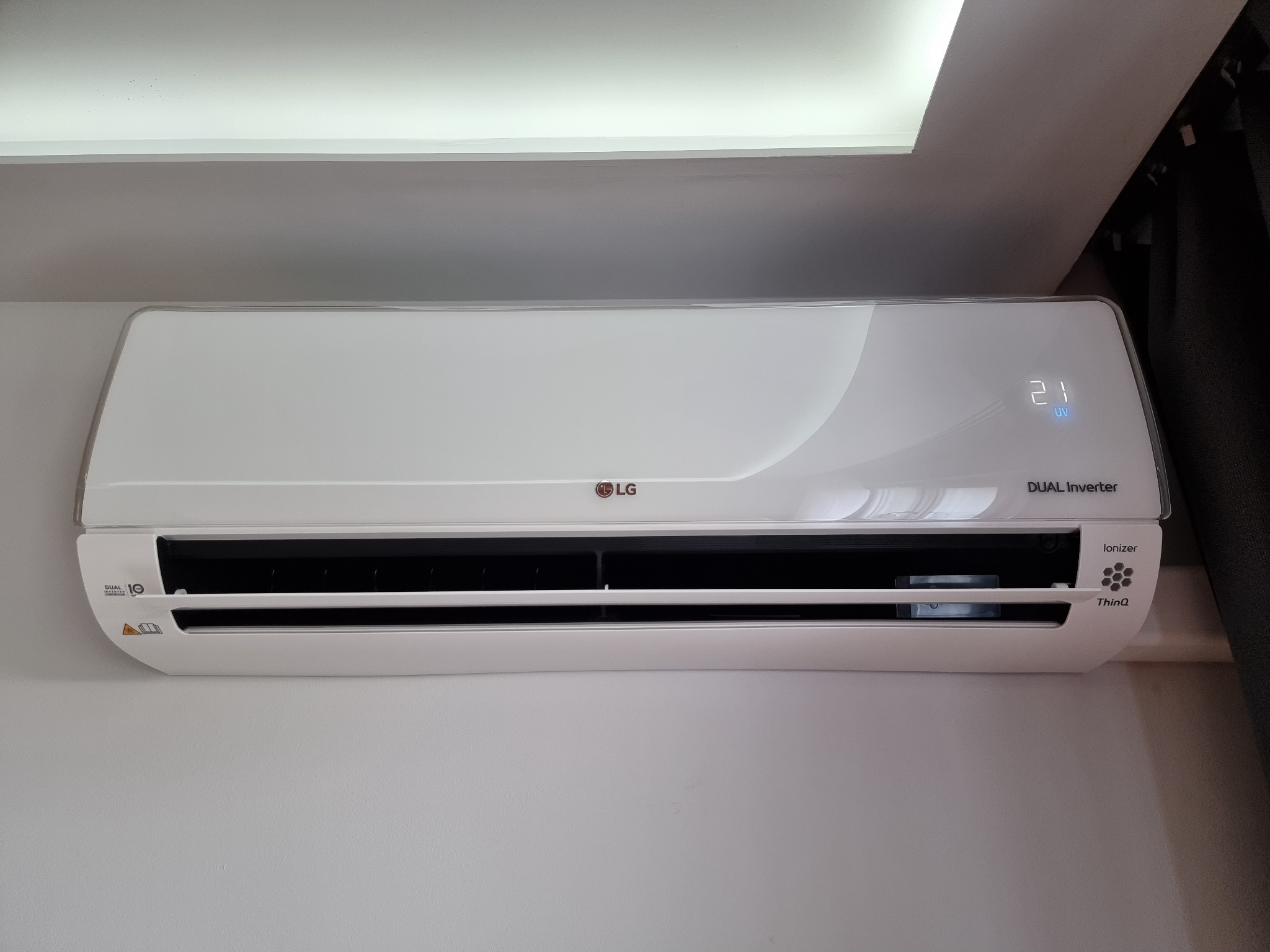 Biała klimatyzacja LG Dual Inverter zamontowana na ścianie, wyświetlacz pokazuje temperaturę 21 stopni, widoczny fragment sufitu z oświetleniem i zasłona w tle.