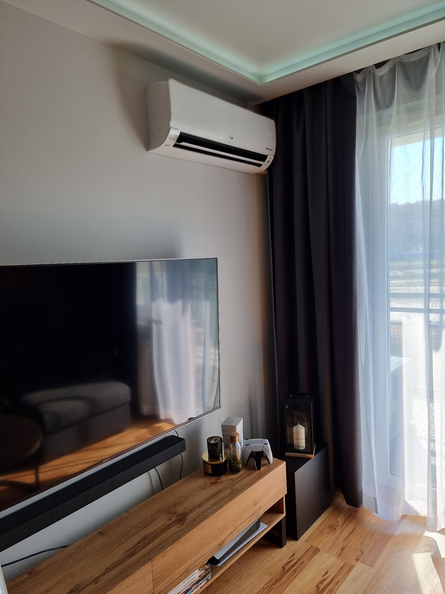 Biały klimatyzator LG zamontowany na ścianie w nowoczesnym salonie z telewizorem, drewnianą komodą, szarymi zasłonami i oknem z widokiem na zewnątrz.