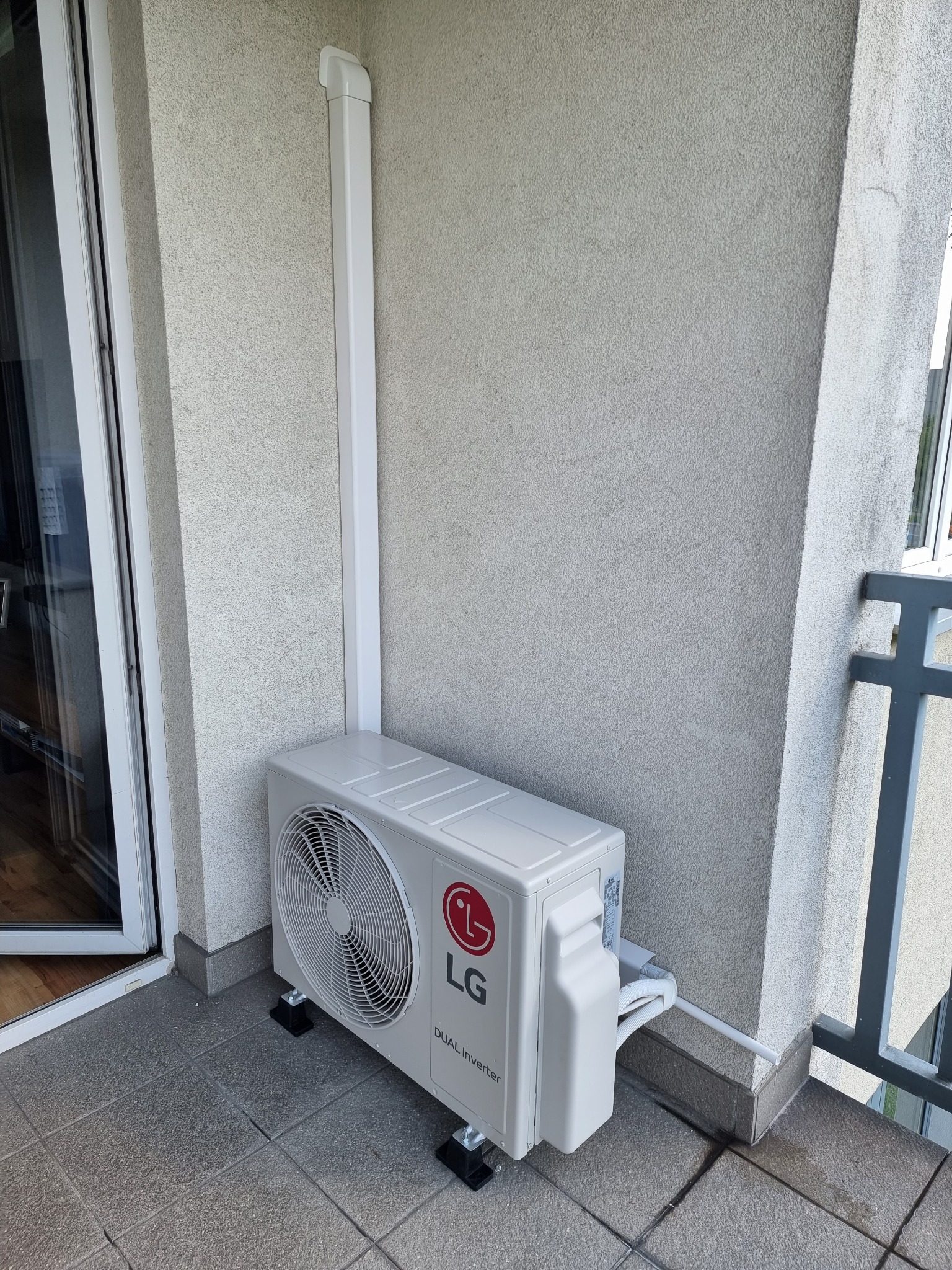 Zewnętrzna jednostka klimatyzacji LG Dual Inverter zamontowana na balkonie, z widocznymi przewodami i rurą odprowadzającą skropliny, przymocowana do ściany budynku o szarej elewacji.