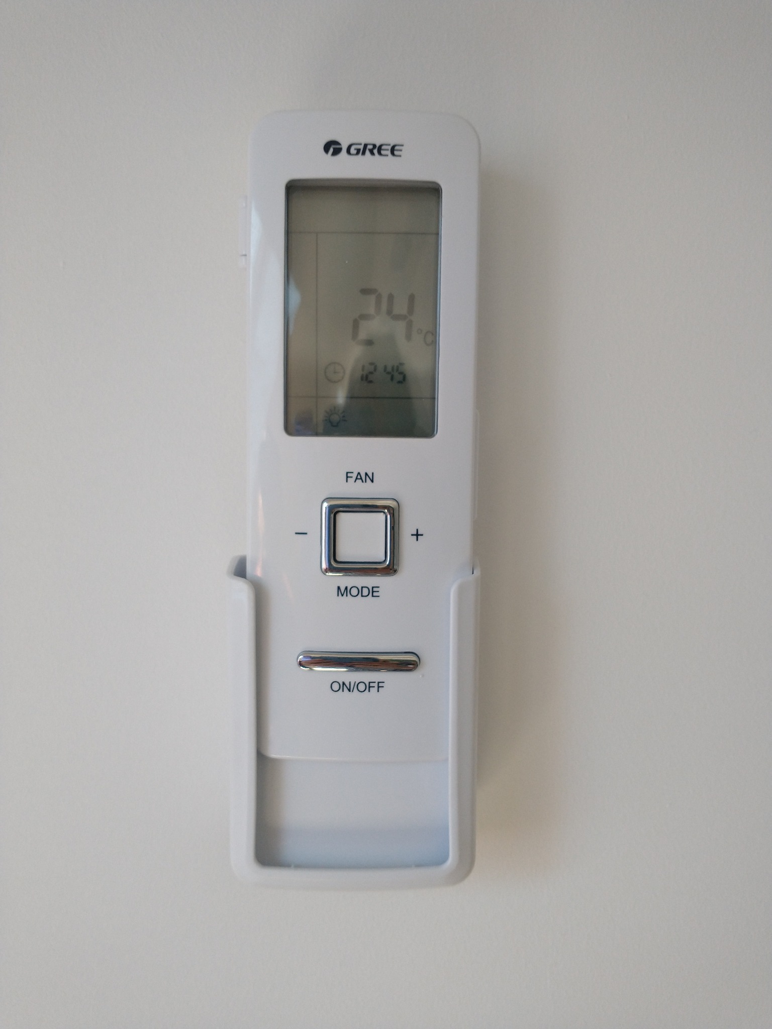 Biały pilot do sterowania klimatyzacją marki Gree, zamocowany w uchwycie na jasnej ścianie, wyświetlający temperaturę 24 stopnie Celsjusza.