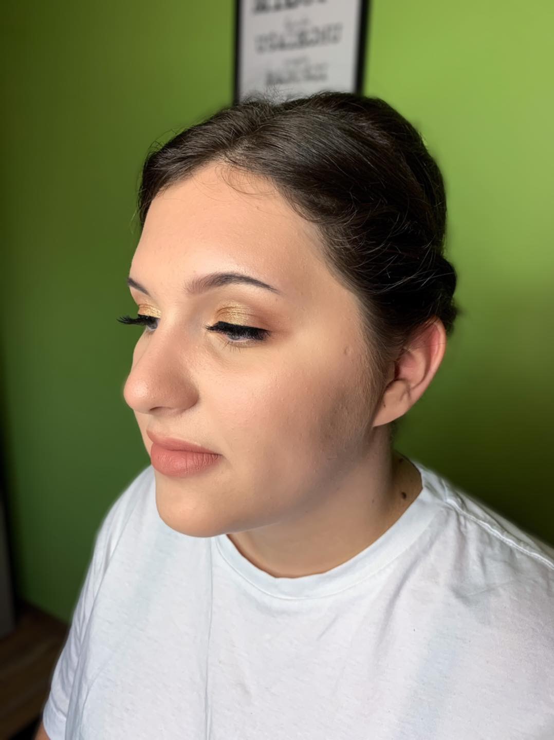 Profil kobiety z makijażem: złote cienie na powiekach, delikatny róż na policzkach i naturalne usta, włosy upięte do góry, zielone tło.