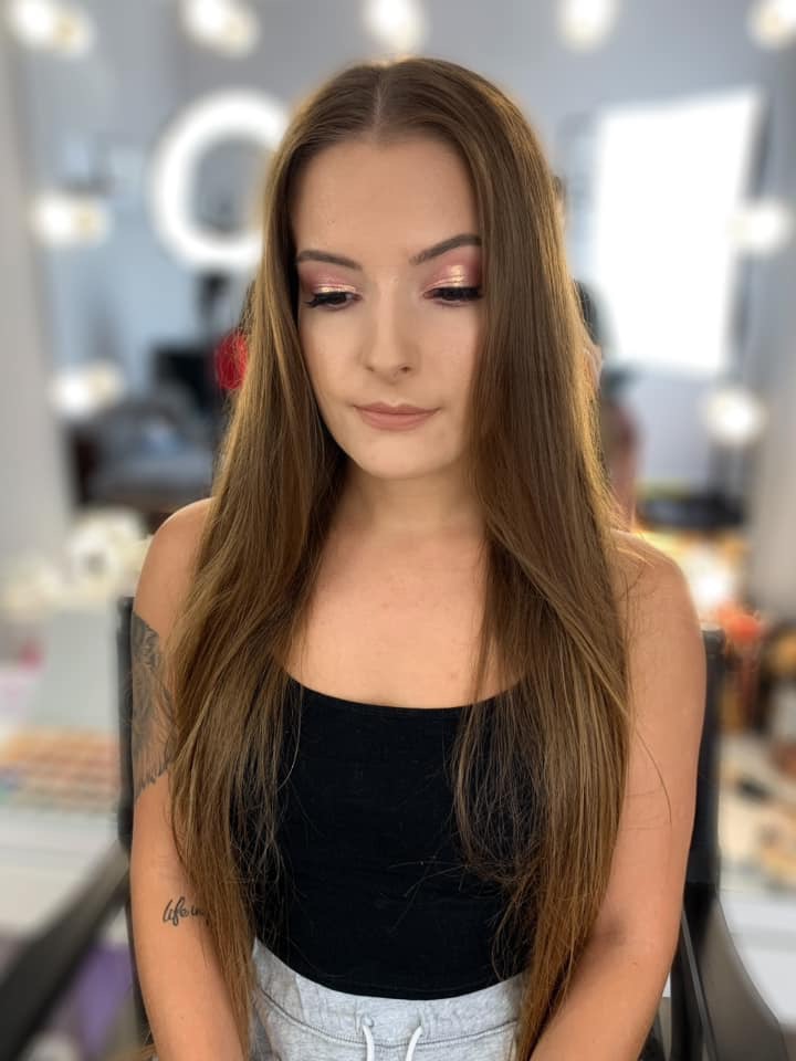 Portret kobiety z błyszczącym, różowym cieniem na powiekach i delikatnym podkreśleniem ust, na tle rozmytego stanowiska wizażysty z lampą pierścieniową.